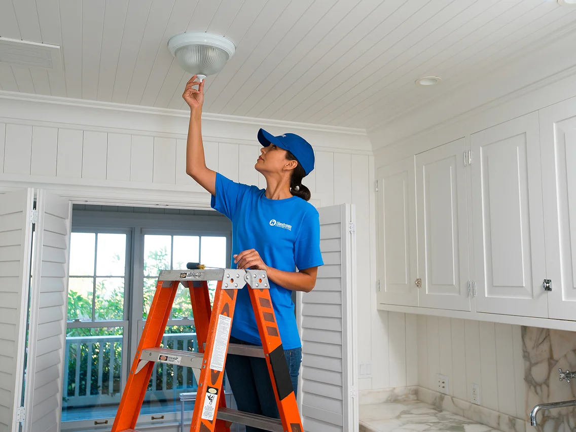 Handyman standing on a ladder installing or replacing a ceiling light fixture inside a home.
