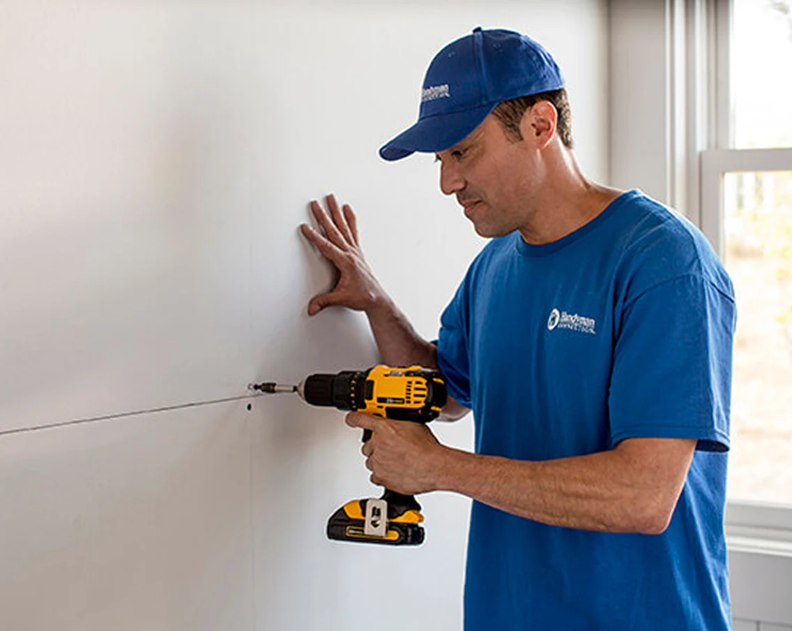 Worker in a Handyman Connection uniform uses a yellow cordless drill to drive a screw into drywall at a seam, bracing the wall with his other hand beside a window.