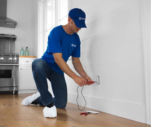 Technician in a blue Handyman Connection uniform kneels on a hardwood floor using a screwdriver to work on an electrical outlet, with tools and wire nearby.