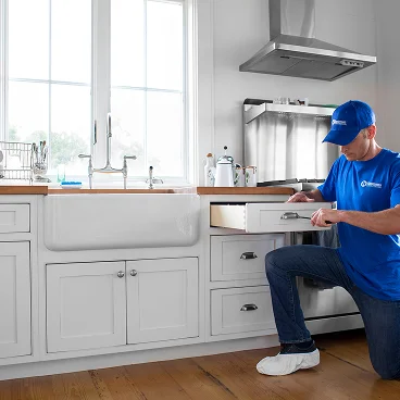 Home service professional kneeling in a kitchen while repairing or adjusting a cabinet drawer.