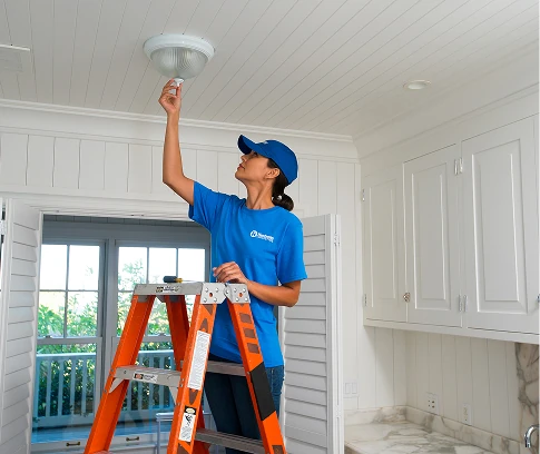 Handyman Connection technician in a blue uniform stands on an orange ladder in a bright kitchen, reaching up to change a lightbulb in a ceiling fixture.