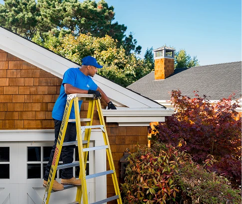 Technician in a blue Handyman Connection uniform stands on a yellow ladder cleaning a house gutter along a shingled roof, with trees and a clear blue sky behind.