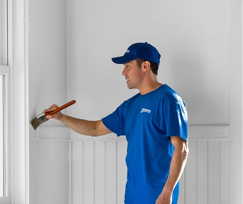 Technician in a blue Handyman Connection uniform paints trim beside a window with a wide brush in a bright room.
