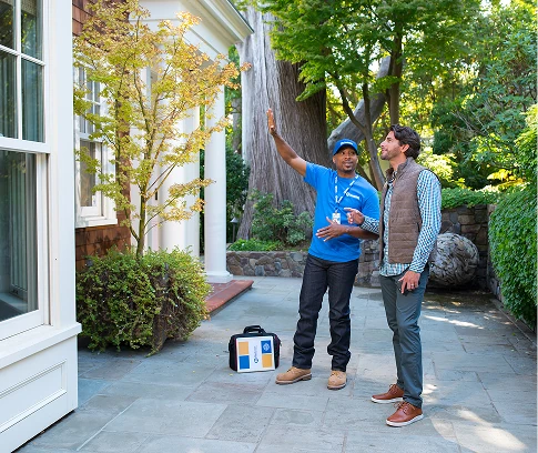 Technician in a Handyman Connection uniform talks with a homeowner outdoors, gesturing toward the house while a tool case sits on the patio.